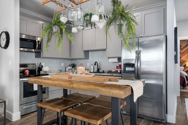 A modern kitchen with stainless steel appliances, a wooden table with benches, hanging plants, and a clock on the wall.