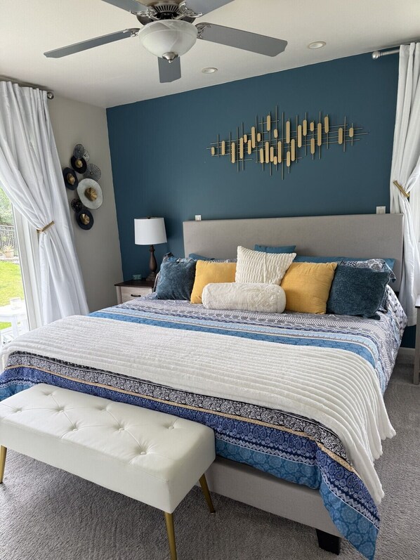 A bedroom with a bed, a ceiling fan, a wall art piece, a bedside table with a lamp, and a window with curtains.