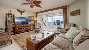 A living room with a large window, a ceiling fan, wicker furniture, and a television.