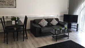 A modern living room with a black leather sofa, two black and white patterned pillows, a black coffee table, and a TV. There is a dining table with chairs and a bottle of champagne on it. A sign on the wall reads "WHEN IT RAINS, LOOK FOR STARS". - Captivating 1-bed Apartment in Swansea (Swansea)
