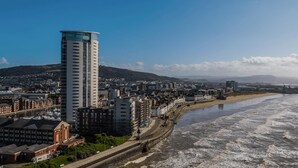 A coastal cityscape with a prominent modern high-rise building, a beach, and a railway track running parallel to the waterfront. - Captivating 1-bed Apartment in Swansea (Swansea)