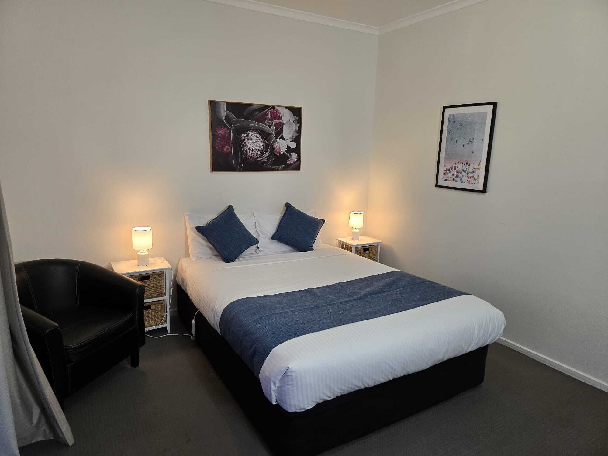 Habitación de hotel ordenada con una cama grande, dos mesitas de noche con lámparas, una silla negra y dos cuadros enmarcados en las paredes.