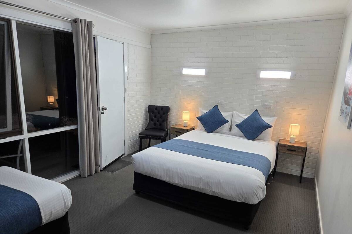 A hotel room with a white brick wall, a large bed with blue bedding, two bedside tables with lamps, a chair, and a sliding glass door.