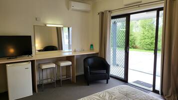 A hotel room with a flat-screen TV, a desk with two bar stools, a black chair, and a sliding glass door leading to a balcony with a view of greenery.