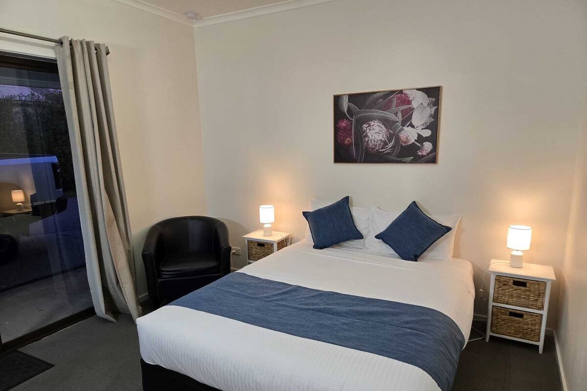 A hotel room with a bed, two bedside tables with lamps, a chair, a window with curtains, and a wall-mounted artwork.