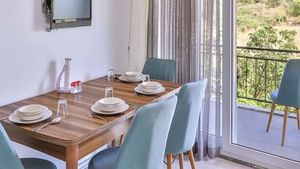 A dining area with a wooden table set for four, a television mounted on the wall, and a view of a balcony with a railing. - Kaya Apart  (Kas)