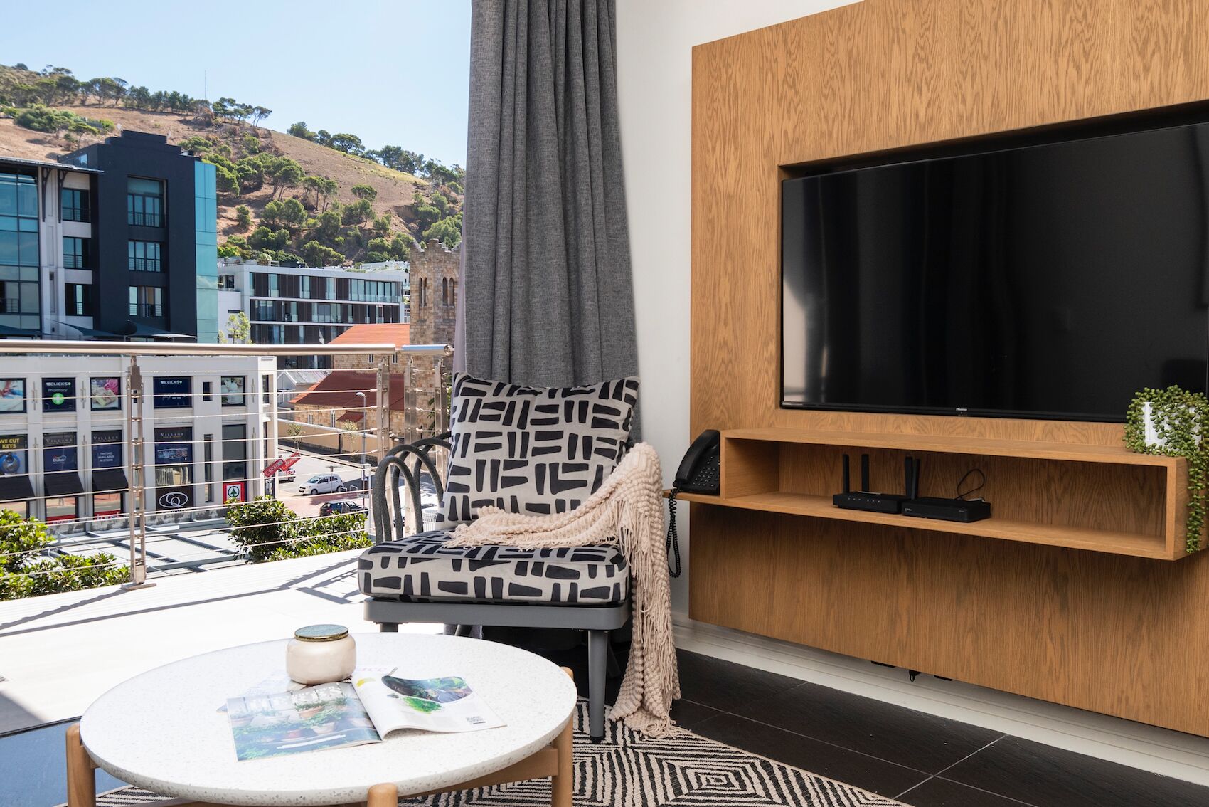 A modern living room with a large TV, a patterned armchair, a small table with magazines, and a view of buildings and hills.