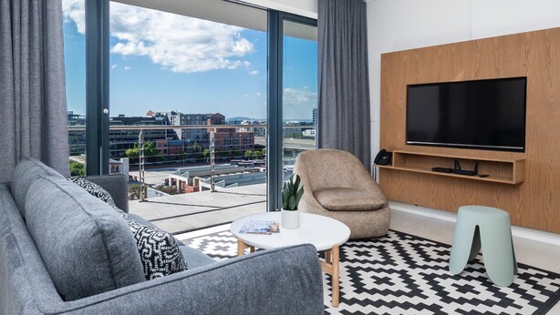 Een moderne woonkamer met een groot raam, een grijze bank, fauteuil en een flatscreen televisie aan de muur.