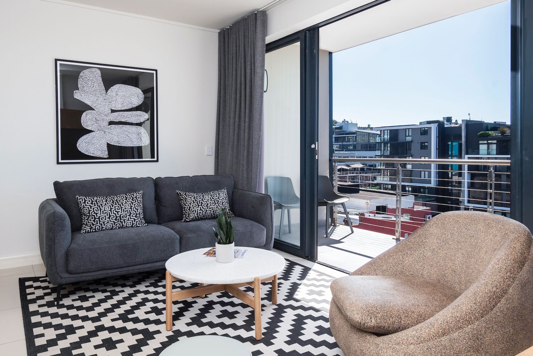 A modern living room with a grey sofa, patterned cushions, a white coffee table, and a brown armchair. A large abstract artwork is on the wall, and the room has a large window with a city view.