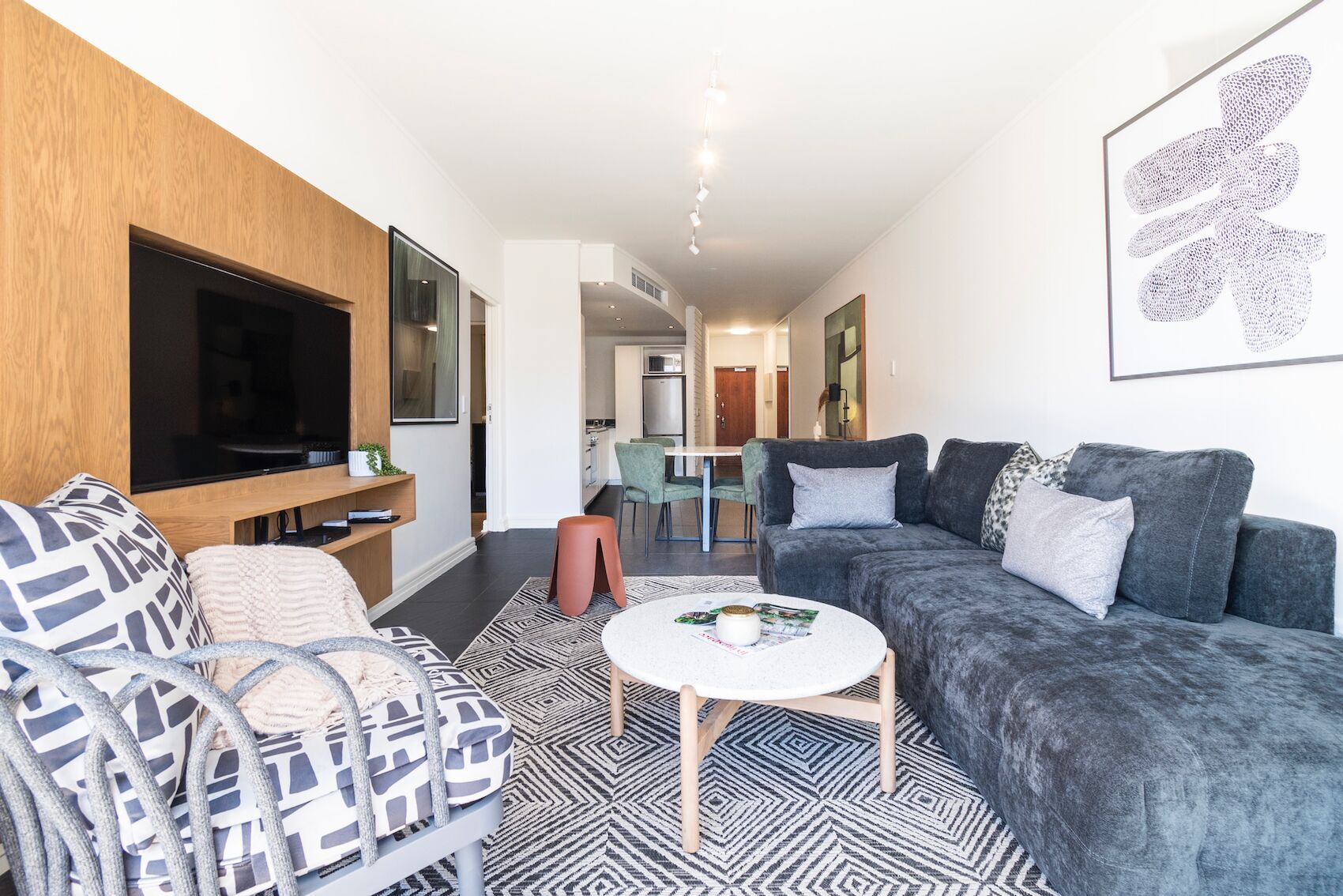 A modern living room with a grey sofa, patterned armchair, and a round coffee table. A flat-screen TV is mounted on the wall, and a dining area is visible in the background.