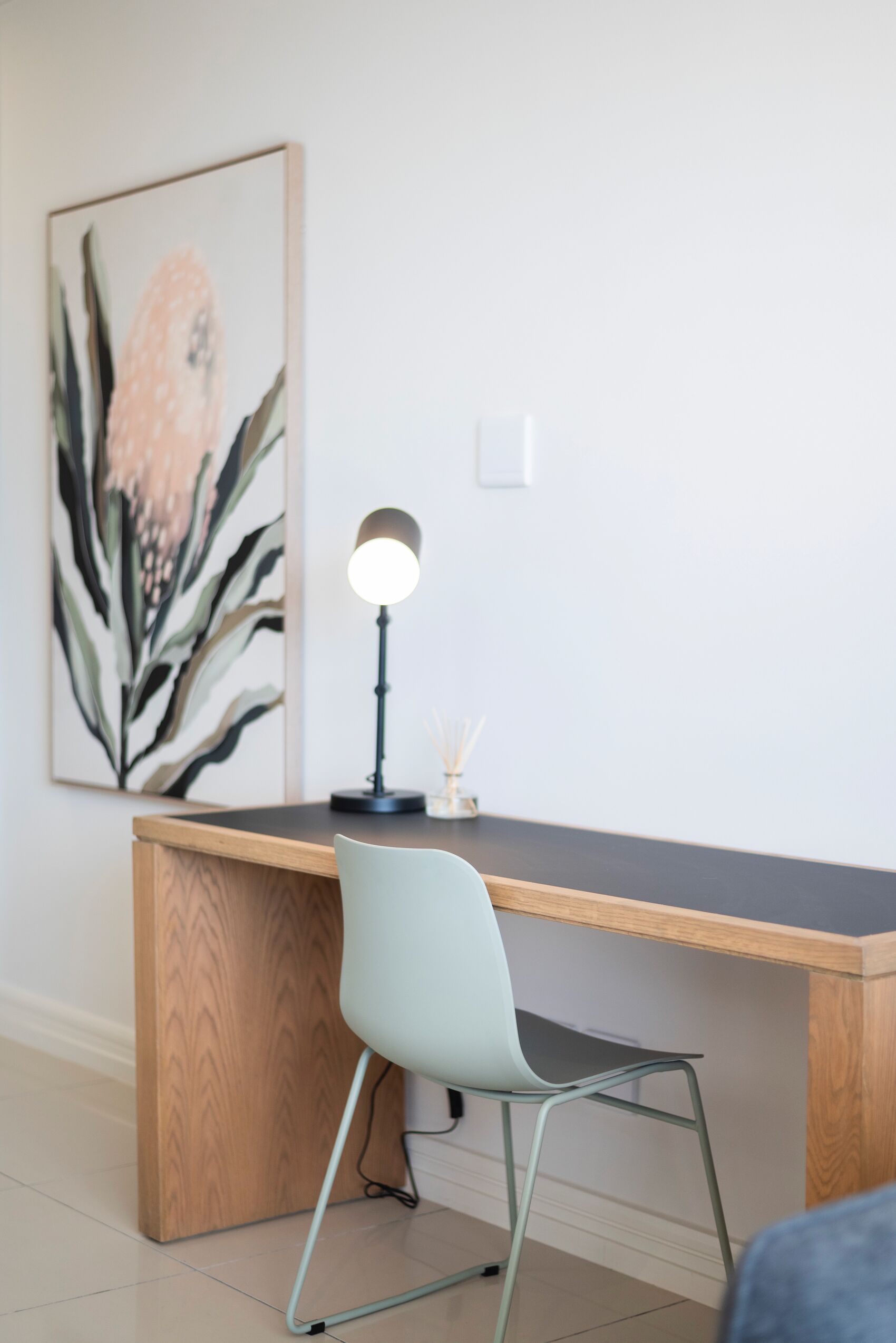 A modern desk with a light-colored chair, a desk lamp, and a framed artwork on the wall.