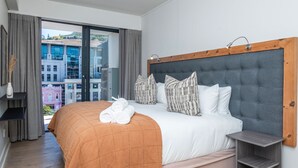 Hotelkamer met een groot bed, uitzicht op gebouwen door een groot raam, een houten hoofdbord en een klein nachtkastje.
