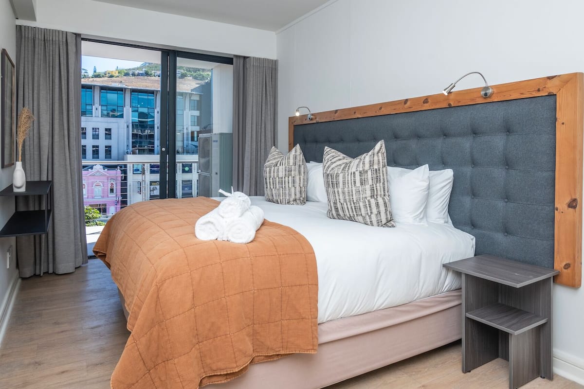 Hotelkamer met een groot bed, uitzicht op gebouwen door een groot raam, een houten hoofdbord en een klein nachtkastje.