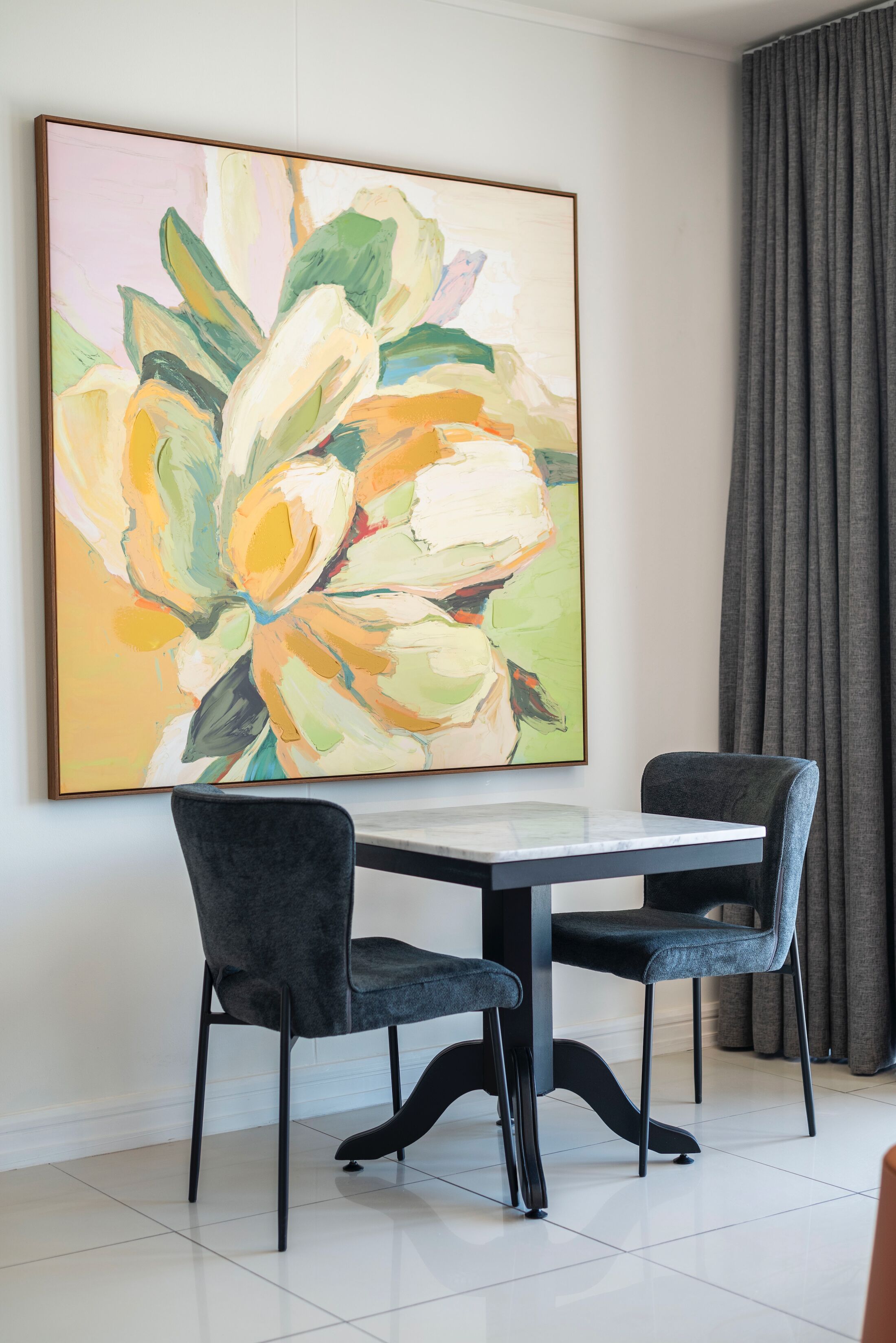 Uma área de refeições com uma mesa e duas cadeiras, uma pintura abstrata grande na parede e uma cortina cinzenta.