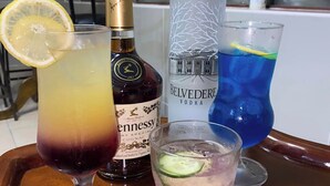 A tray with three drinks: a gradient cocktail, a bottle of Cognac, and a blue drink with a lemon slice. - Dikukama Hotel (Tshabong)