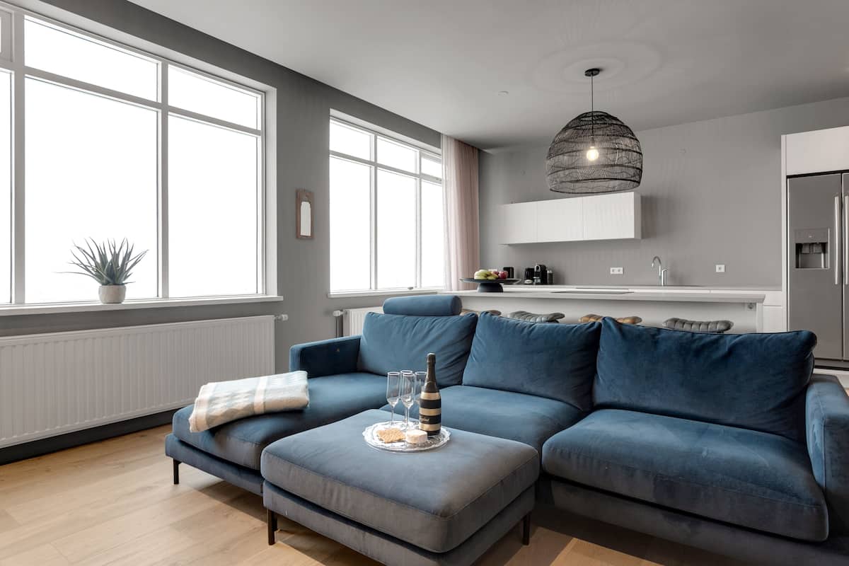 A modern living room with a grey sectional sofa, a small ottoman, and a dining table set with glasses and a bottle.