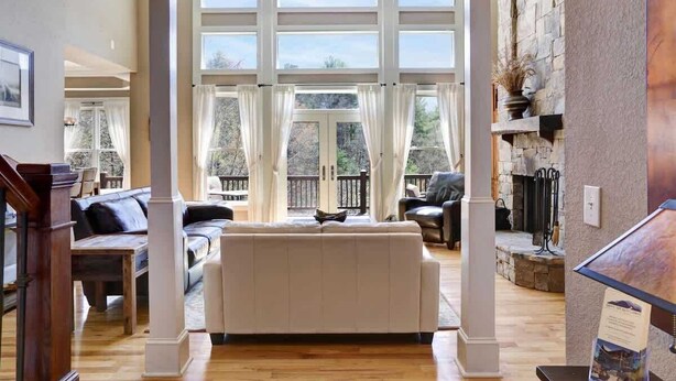 Een ruime woonkamer met een grote witte bank, houten vloeren en een uitzicht op de omgeving door grote ramen.