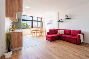 Uma sala de estar moderna com um sofá vermelho, piso de madeira, área de jantar com cadeiras e cozinha com pia e planta em vaso.