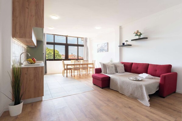 Uma sala de estar moderna com um sofá vermelho, uma mesa de jantar com cadeiras, uma área de cozinha com uma pia e uma planta em vaso, e uma janela grande com vista para o exterior.