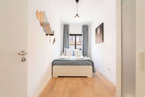 Une chambre à coucher avec un lit, des rideaux, un luminaire fixé au mur et une fenêtre donnant sur des immeubles.