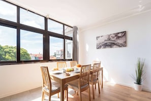 Uma área de refeições com mesa e cadeiras de madeira, uma planta em vaso e vista para edifícios através de amplas janelas.