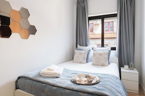 Uma cama bem arrumada com colcha azul estampada, um suporte com itens de café da manhã e uma luminária hexagonal fixada na parede.
