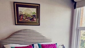 Ein ordentlich bezogenes Bett mit gestreifter Bettwäsche und farbenfrohen Kissen, ein gerahmtes Bild an der Wand und ein Fenster mit Blick auf Grünflächen.