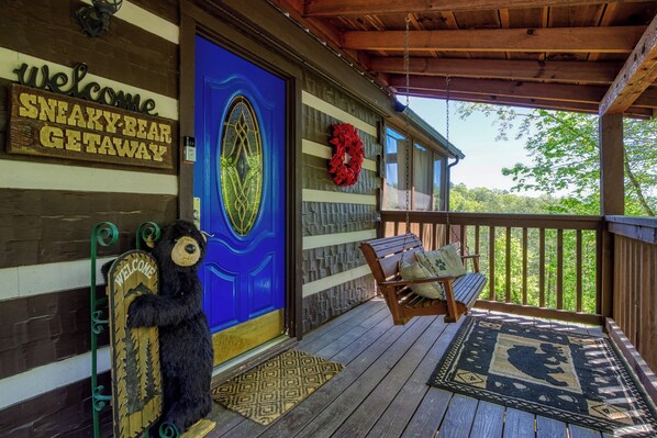 A wooden porch with a blue door, a welcome sign, a bear decoration, and a hanging chair.
