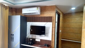 A modern room with a built-in air conditioner, a flat-screen TV, a wooden vanity, and a small seating area with cushioned stools. - Hotel Asha Delight (Narendranagar)