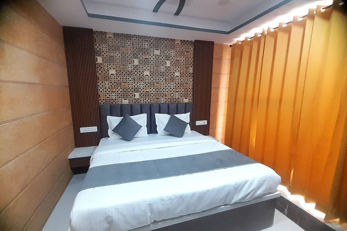 A neatly made bed with a headboard, a wall with a decorative panel, and a window with blinds.