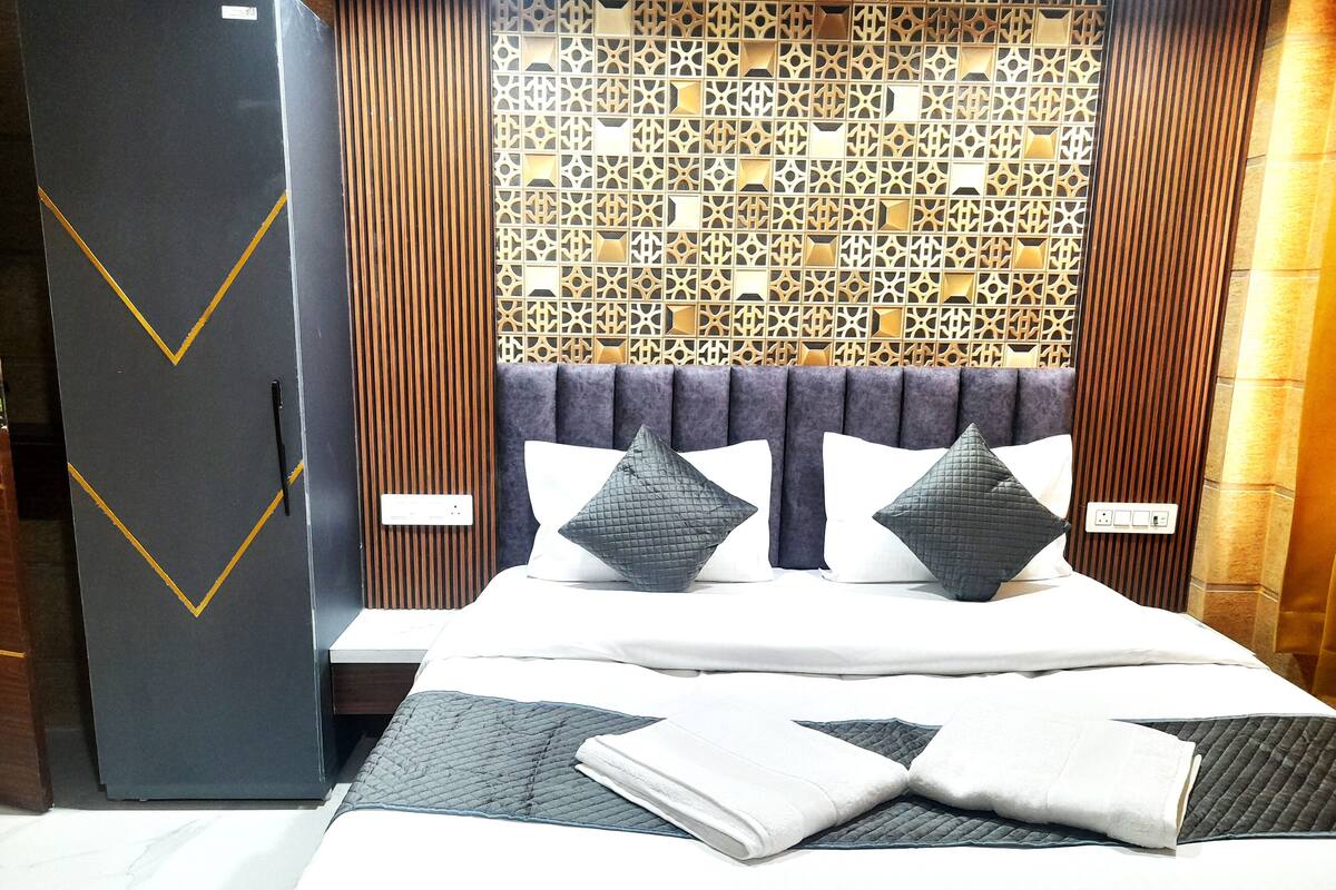A neatly made bed with pillows and a folded towel, a wooden panel wall with a geometric pattern, and a dark-colored wardrobe with a gold stripe.