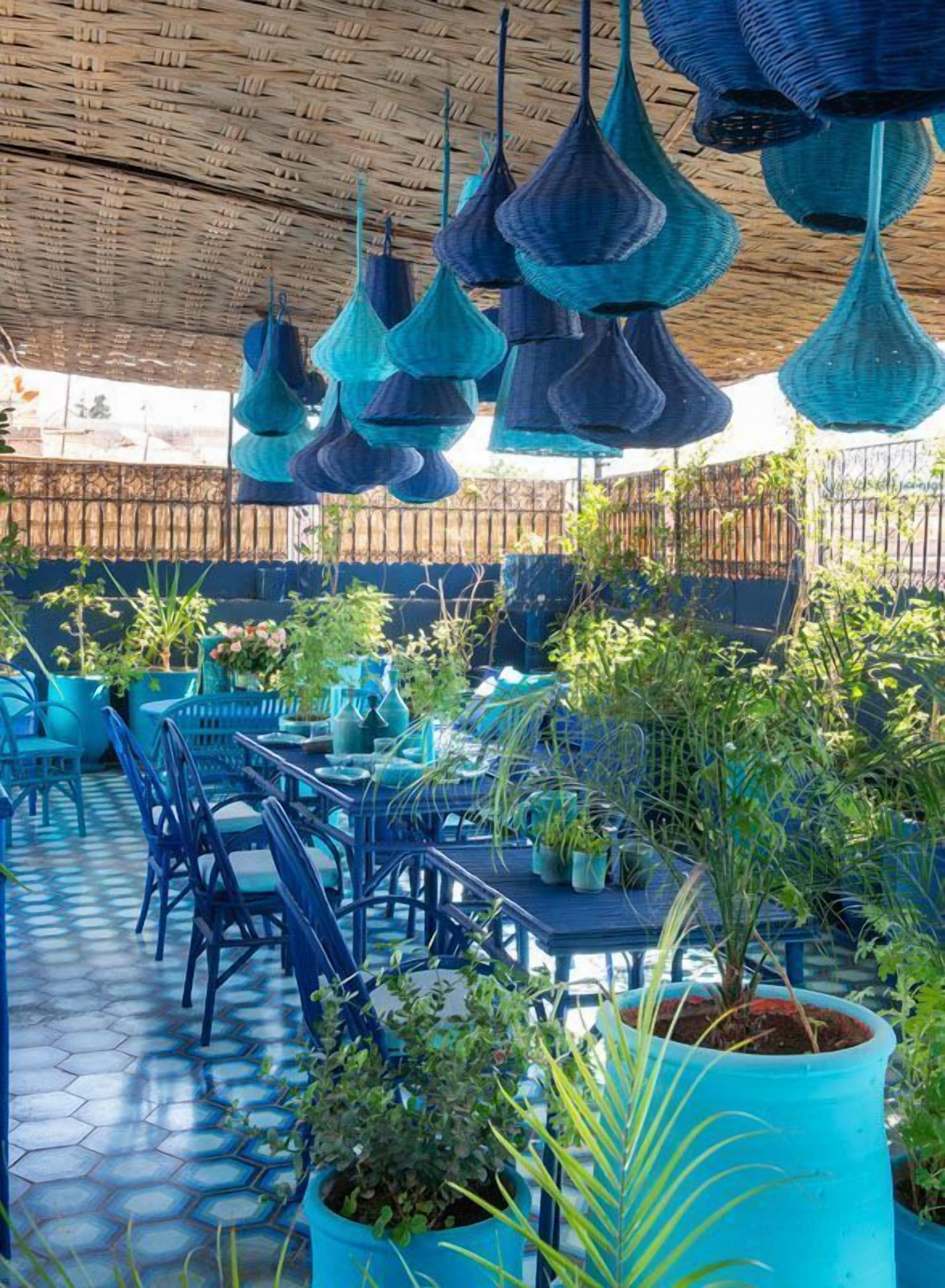 Un área de estar al aire libre con sillas y mesas azules, rodeada de plantas en macetas y lámparas azules colgantes.