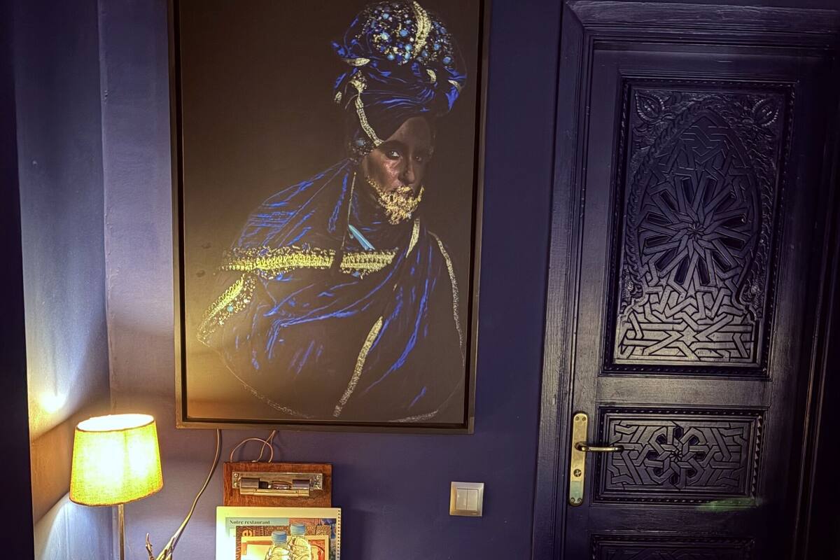 Una habitación con una puerta azul oscuro, una obra enmarcada en la pared, una mesa de madera con una lámpara, una silla y una bandeja con vasos y un teléfono.