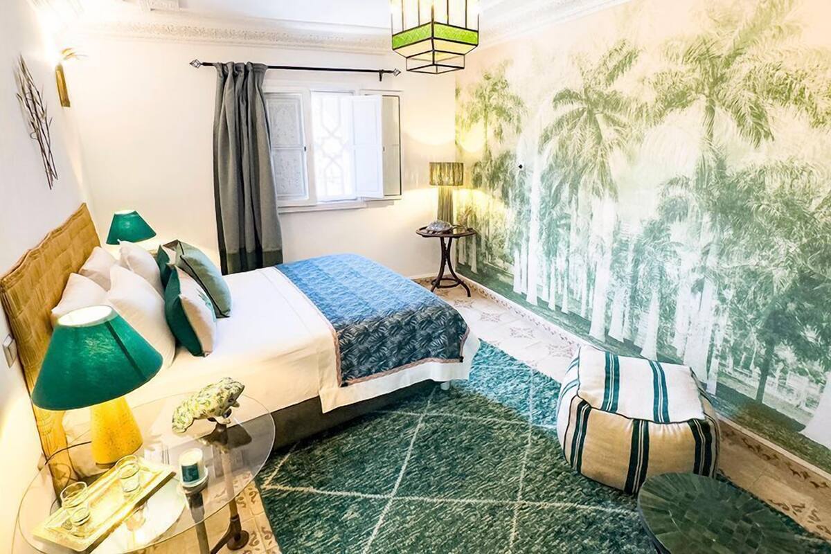 Habitación de hotel con cama, una mesita de noche con lámpara, un mural decorativo y una ventana con cortinas.