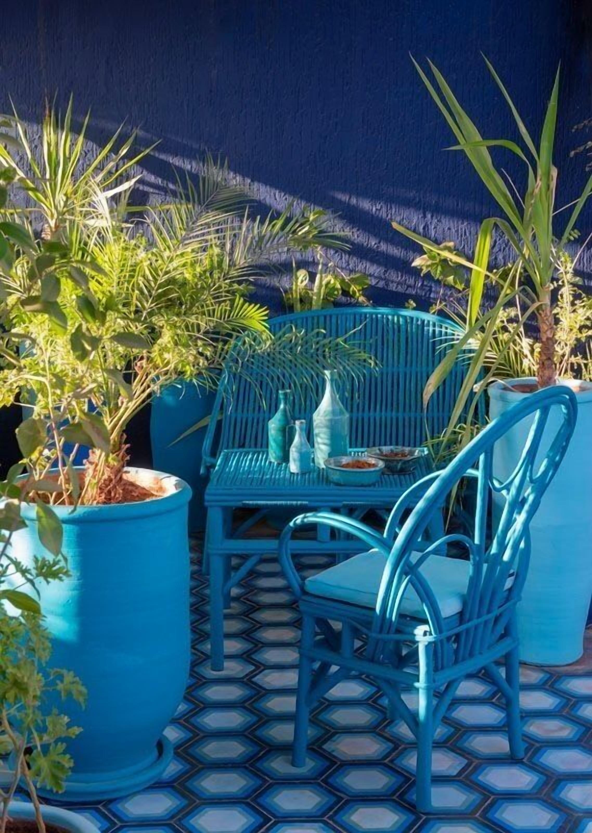 Un espacio al aire libre con asientos azules, una mesa y sillas, rodeado de plantas en macetas.