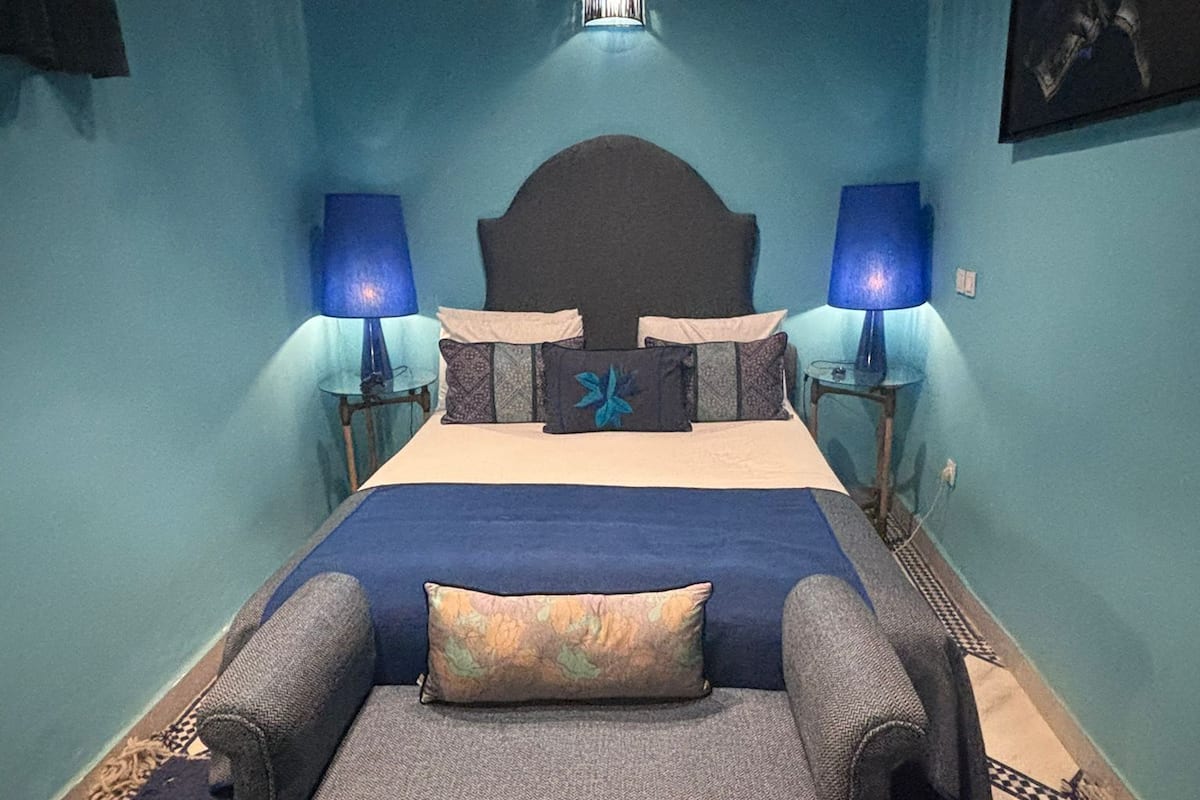 Un dormitorio con una cama, dos mesitas de noche con lámparas, un banco al pie de la cama y una lámpara de pared.
