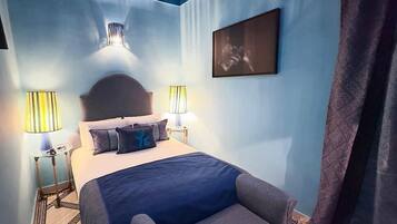 A bedroom with a bed, two bedside lamps, a headboard, a nightstand, a chair, a painting on the wall, and a patterned curtain.