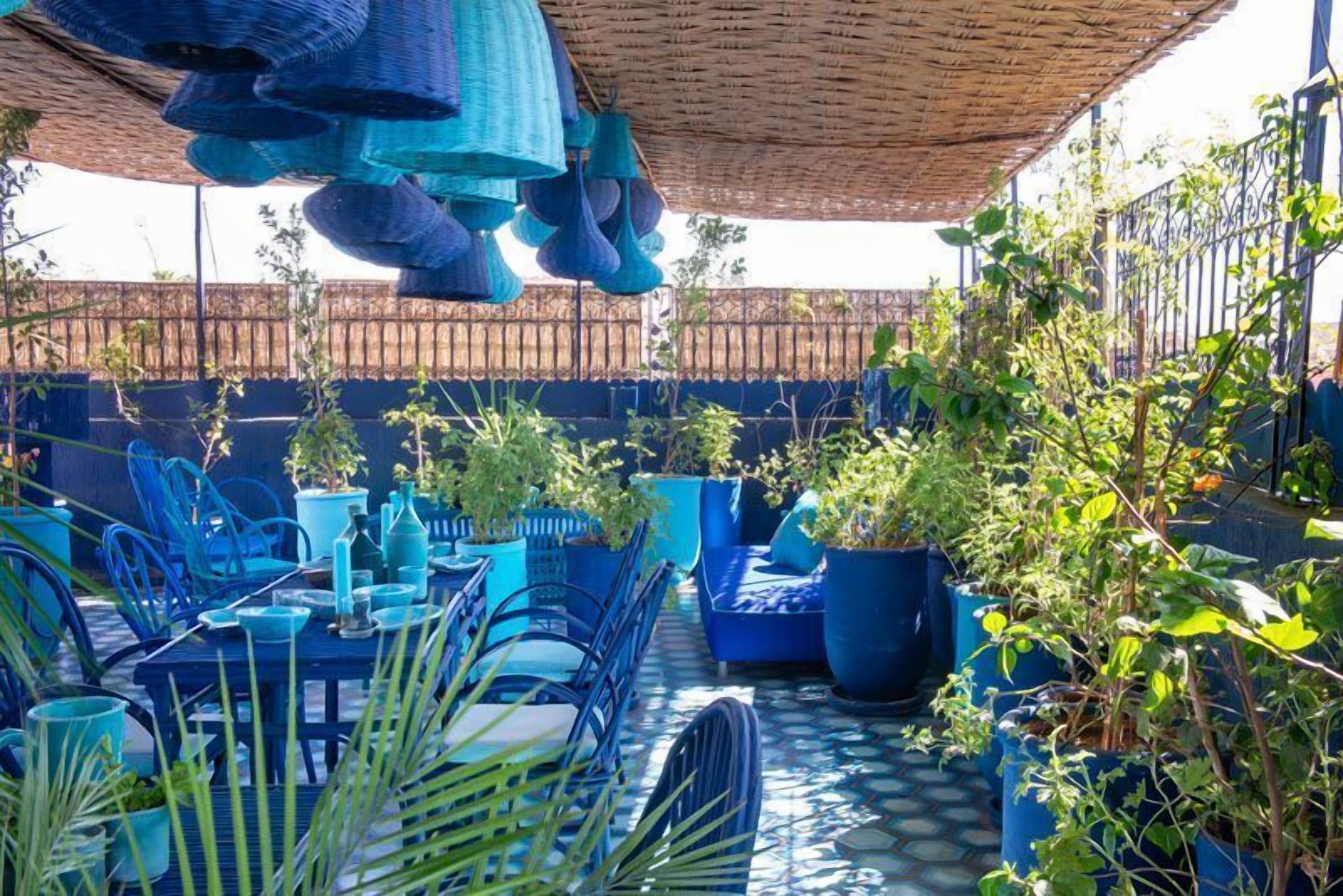 Een terras buiten met blauw meubilair, plantenpotten en een rieten dak.