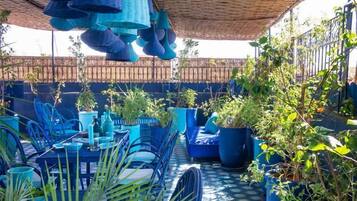 Een terras buiten met blauw meubilair, plantenpotten en een rieten dak.