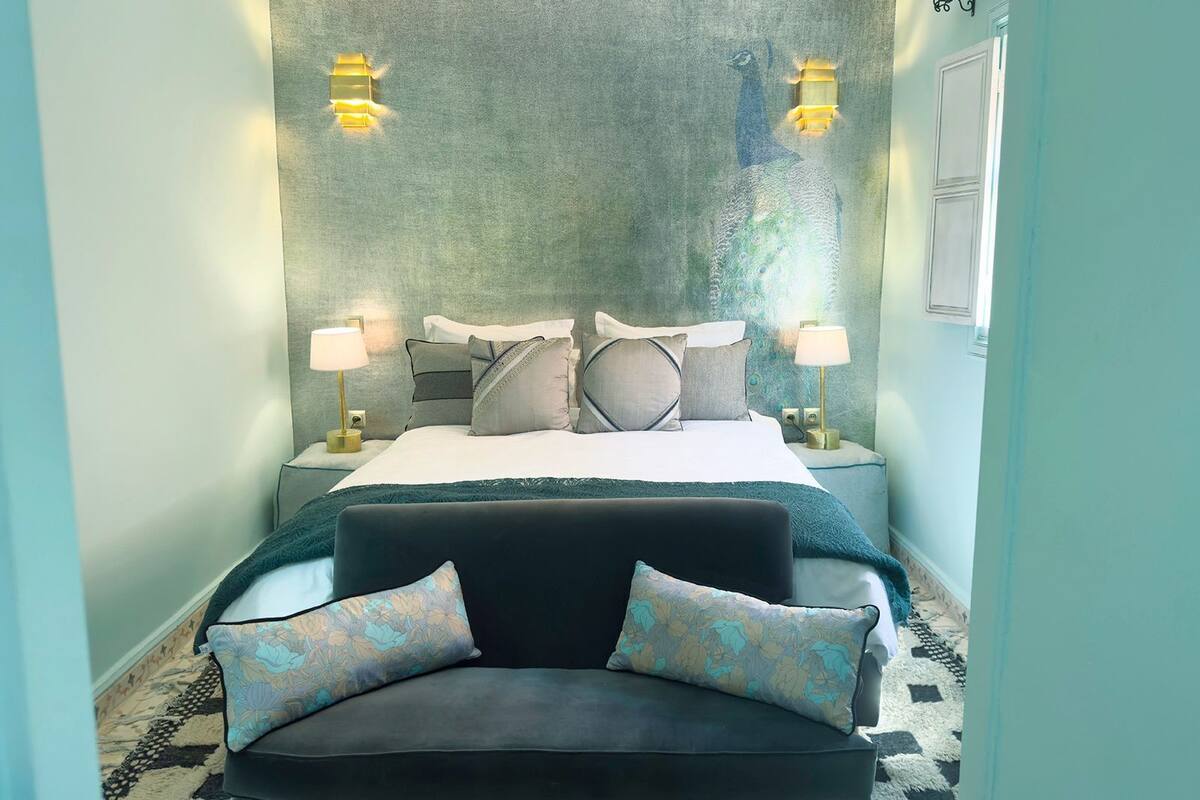 Un dormitorio con cama, dos mesitas de noche con lámparas, un sofá negro con cojines estampados y una pared con textura.