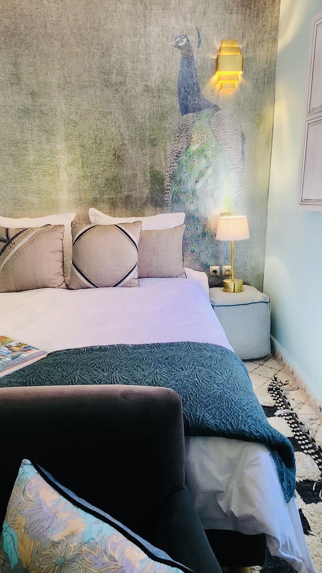 Een slaapkamer met een bed, kussens, een nachtkastje, een lamp en een muurschildering.