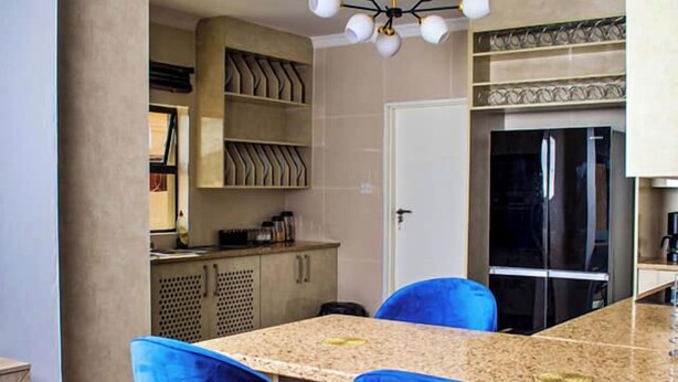 A modern kitchen with a marble countertop, two blue bar stools, a black refrigerator, and a ceiling light fixture. - Ascentia Guest House (Jwaneng)
