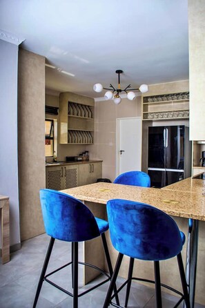 Een moderne keuken met een marmeren aanrecht, twee blauwe barkrukken, een zwarte koelkast en een plafondlamp.