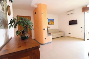 A living room with a wooden bench, a potted plant, a beige sofa, a television mounted on the wall, and a door leading outside. - Calipso's Home in Otranto (Otranto)