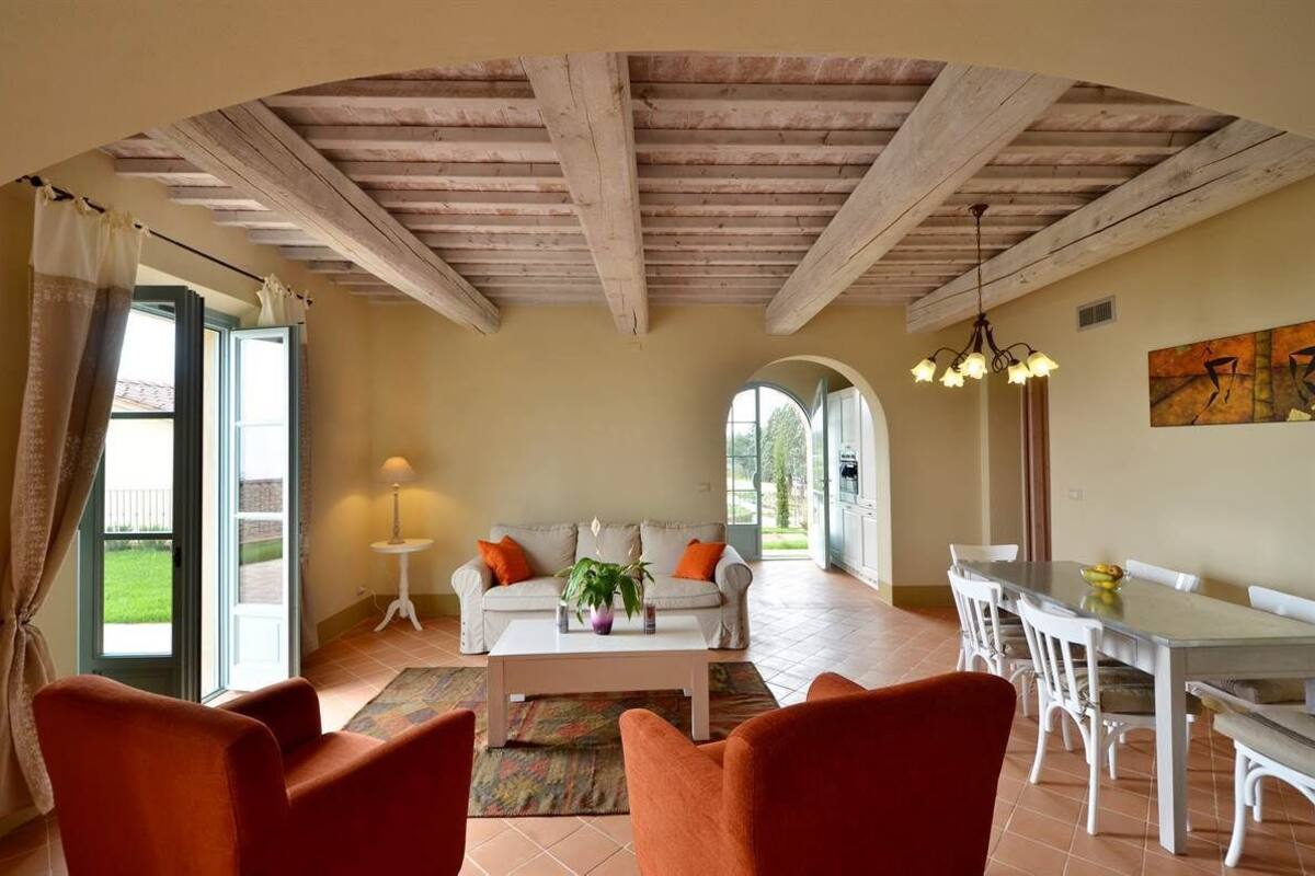A spacious living room with a high wooden ceiling, a white sofa with orange cushions, a coffee table with a plant, and a dining area with white chairs and a table.