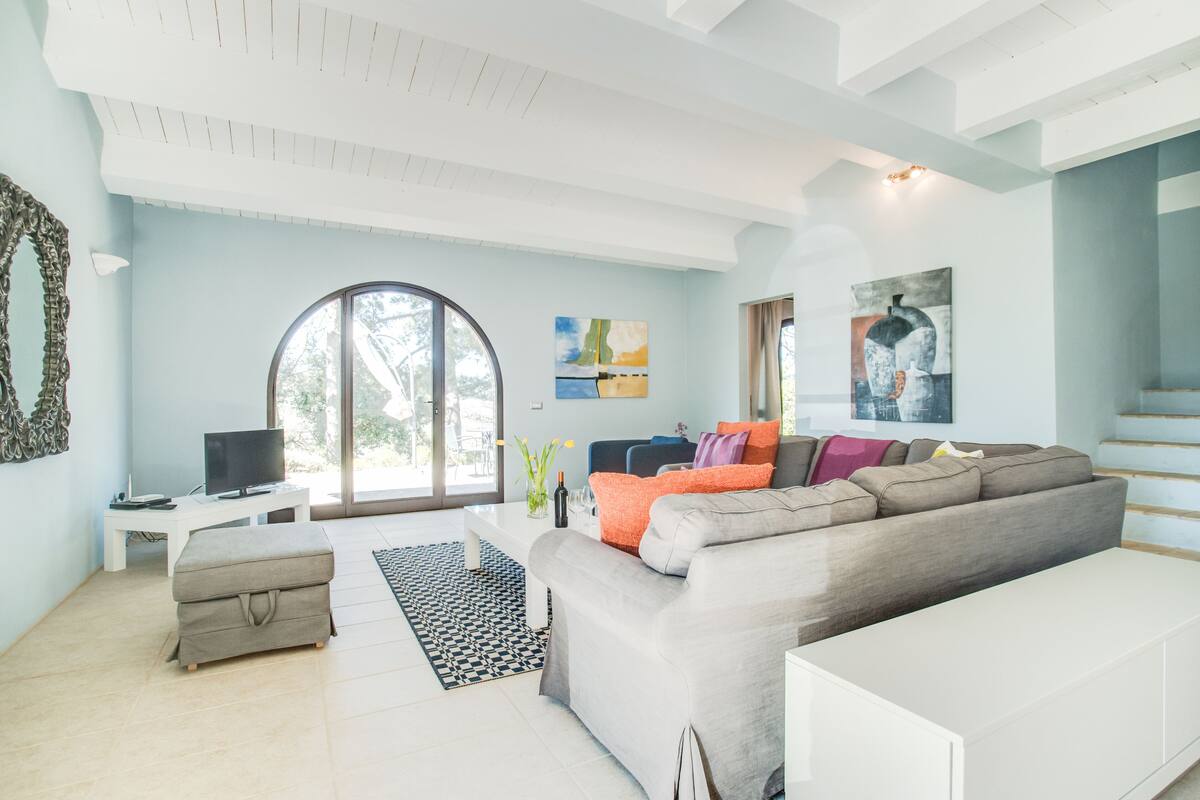A modern living room with a grey sofa, a white coffee table, a TV on a white stand, a large window, and abstract art on the wall.