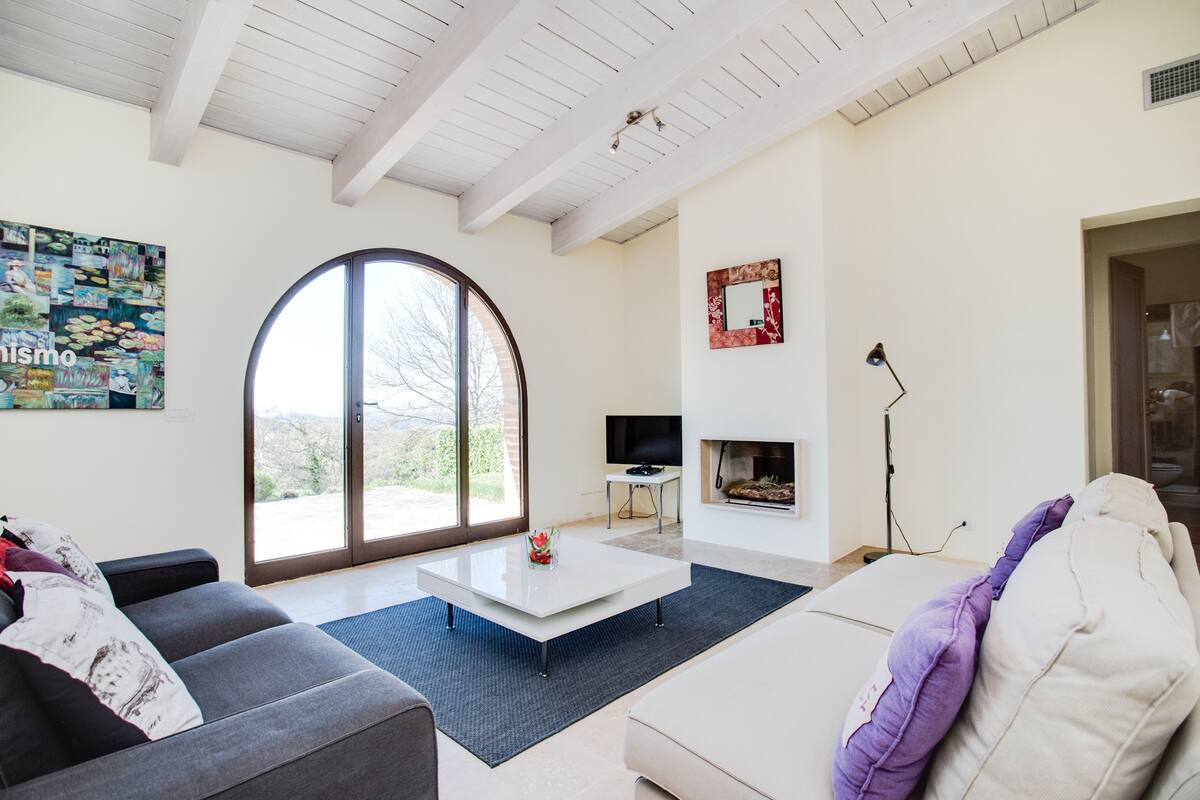 A modern living room with a large arched window, a flat-screen TV, a fireplace, and a cozy seating area.