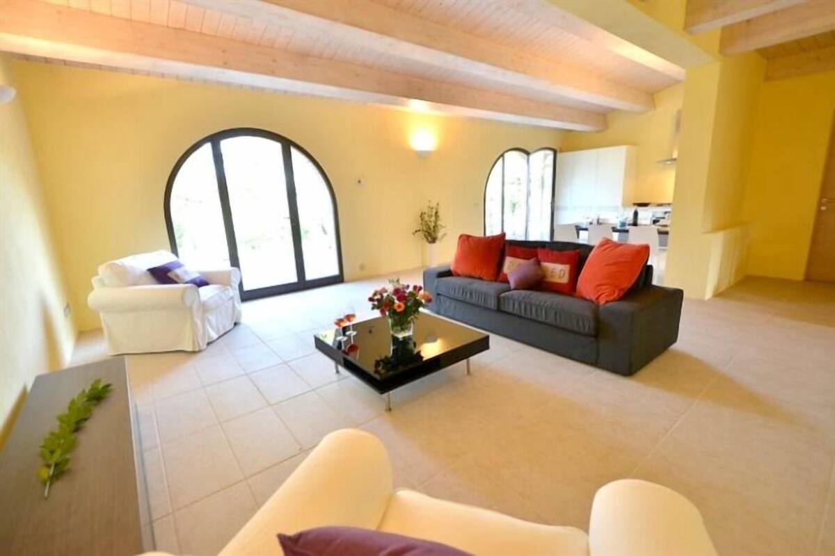 A living room with a sofa, armchairs, a coffee table with flowers, and a large window.