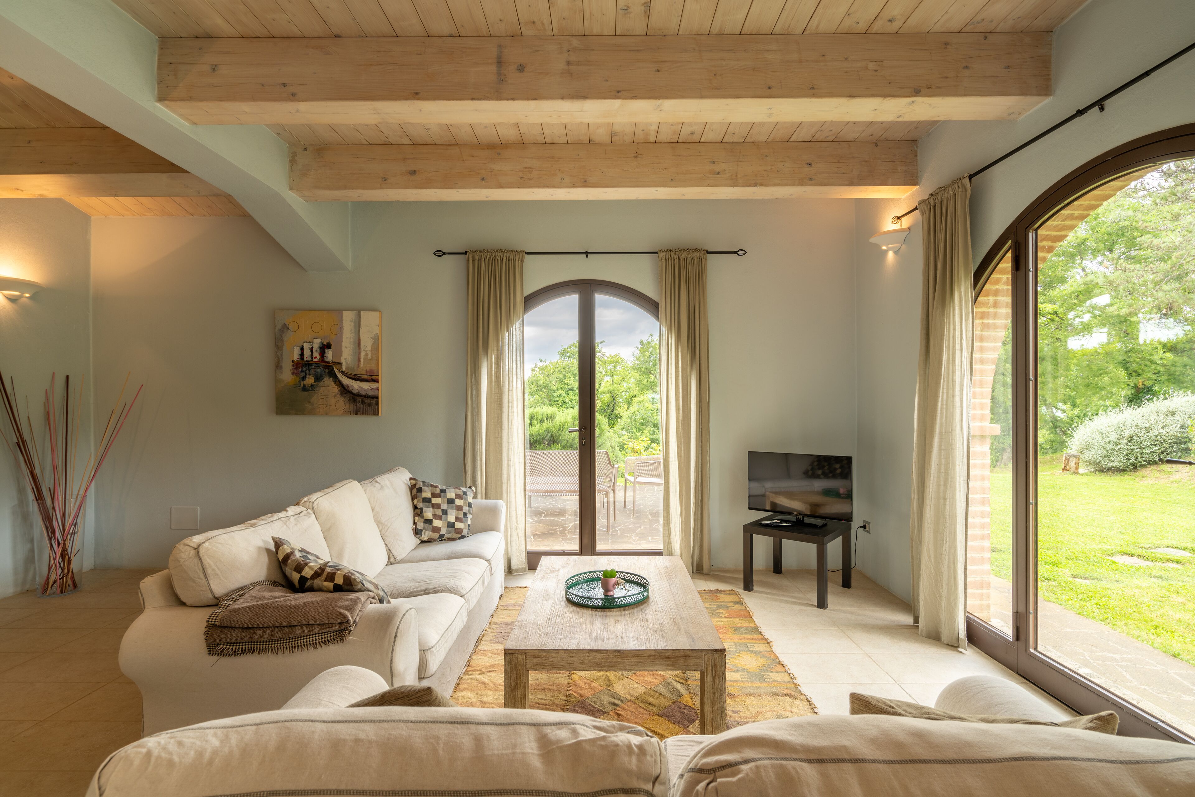 A spacious living room with a wooden ceiling, a large arched window, a sofa, a coffee table, and a TV.