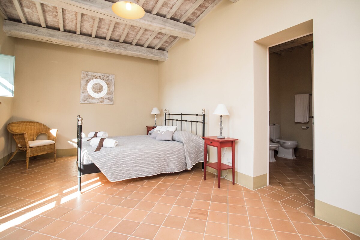 A bedroom with a bed, bedside lamps, a wicker chair, and a view into the bathroom.